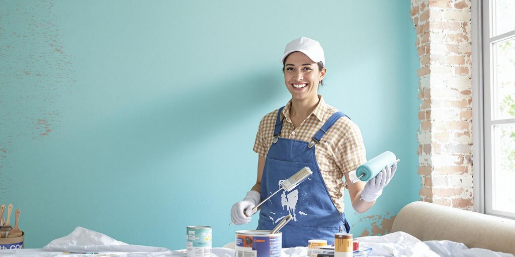 Painter in blue overalls applying turquoise paint to brick wall.